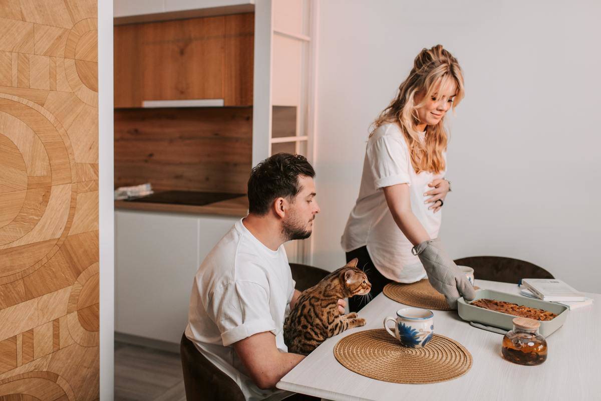 Pregnant woman serving food to man at table with cat on his lap