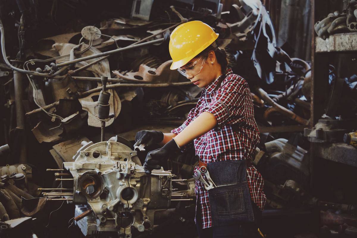 Woman wearing a yellow hard hat, safety goggles, and latex gloves working on motor or engine