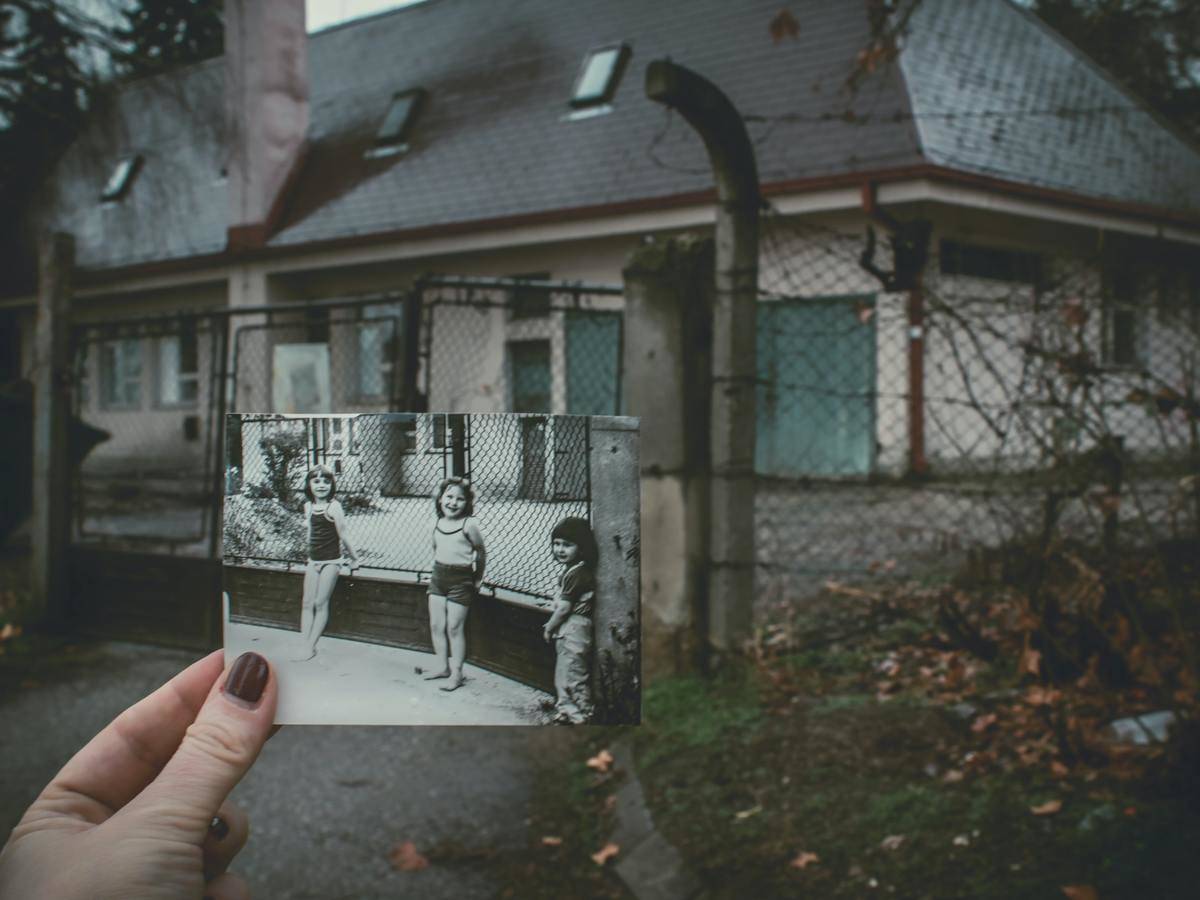 Woman holding up photo of herself as a child in front of her childhood home