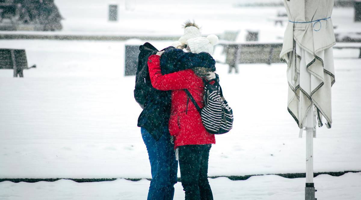 people in winter coats hugging amid snowfall