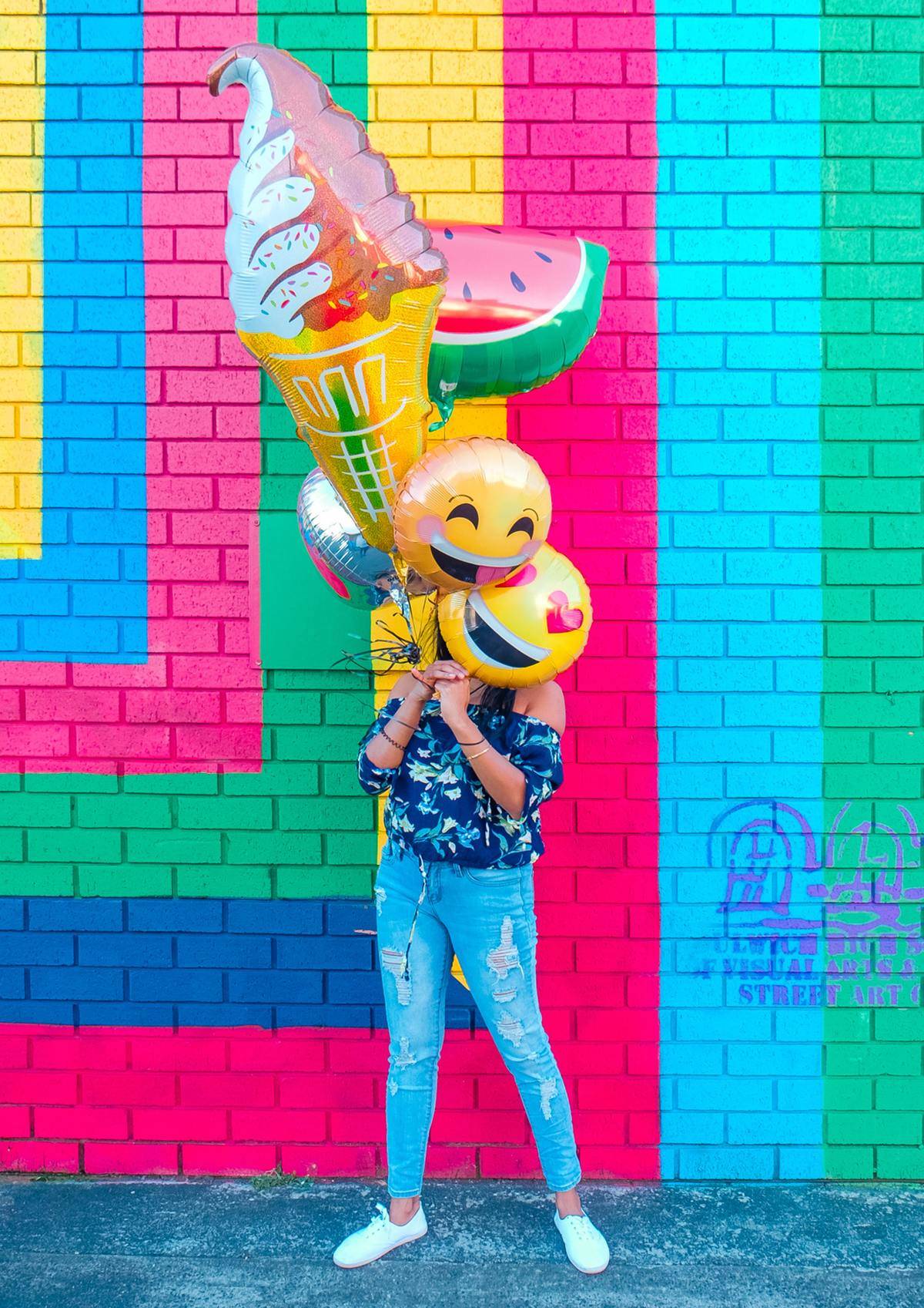woman holding emoji balloons over her face in front of colorful wall