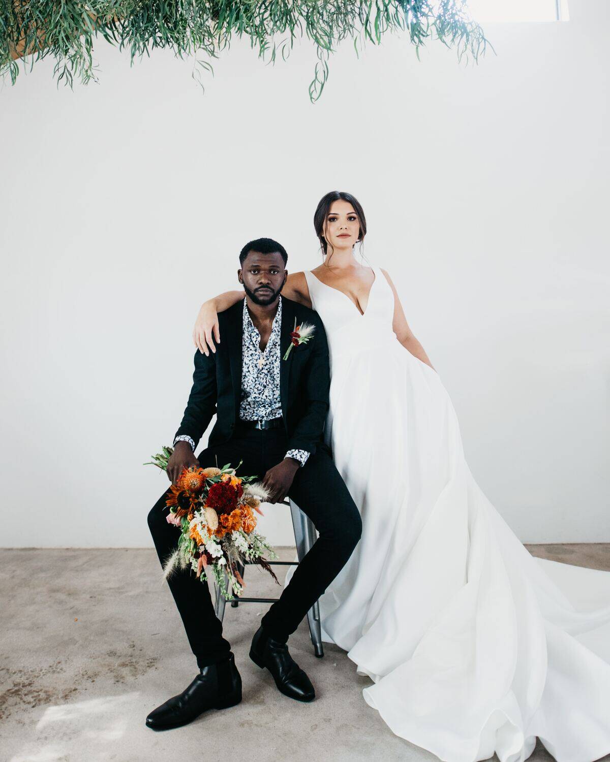 groom sitting and holding flowers with blank expression while bride stands with arm over his shoulder