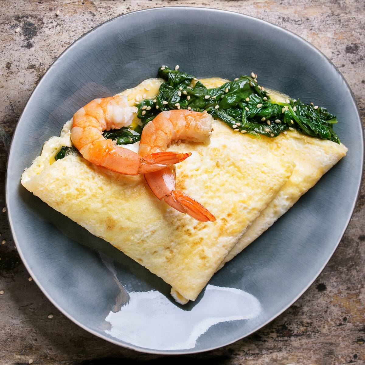 Gray oval plate with omelet staffed cooked spinach and fried shrimps prawns, served with sesame seeds over old iron background. Top view. Square image
