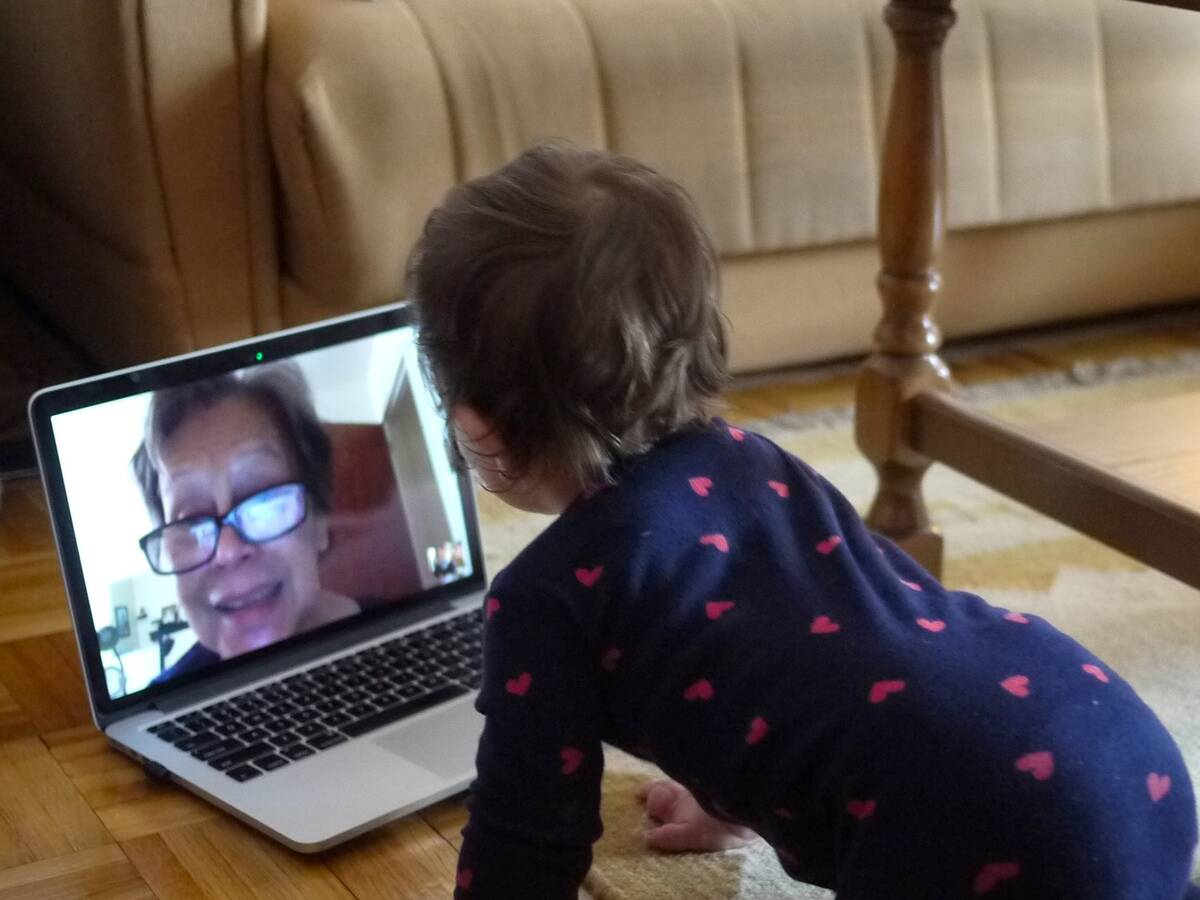 Toddler kneeling before a laptop screen, on a video call with an older female (presumably their grandmother). The scene appears to take place in a living room
