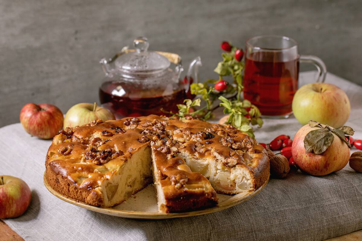 Homemade apple and walnuts cake Charlotte on plate with fresh gardening apples, nuts, rose hip tea around on grey linen table cloth, Autumnal home baking,